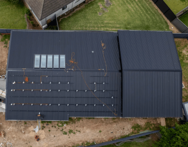 Aerial view of a large dark roof with skylights and solar panel mounts, surrounded by grass and a pathway.