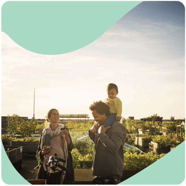 A family enjoys a sunny day on a rooftop garden; a woman with a bike talks to a man carrying a child on his shoulders.