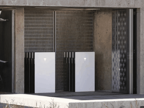 Several Tesla Powerwall battery units are installed against a wall in a concrete enclosure, near a metal fence and sliding door.