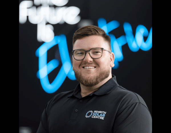 A smiling man in a black polo shirt standing in front of a blurred sign that reads "the future is electric."