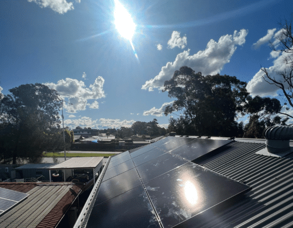 Solar panels installed on a rooftop under a sunny, partly cloudy sky with trees and houses in the background.